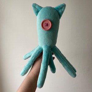 May include: A light blue plush octopus with a pink button for an eye. The octopus has six tentacles and is held in a person's hand.