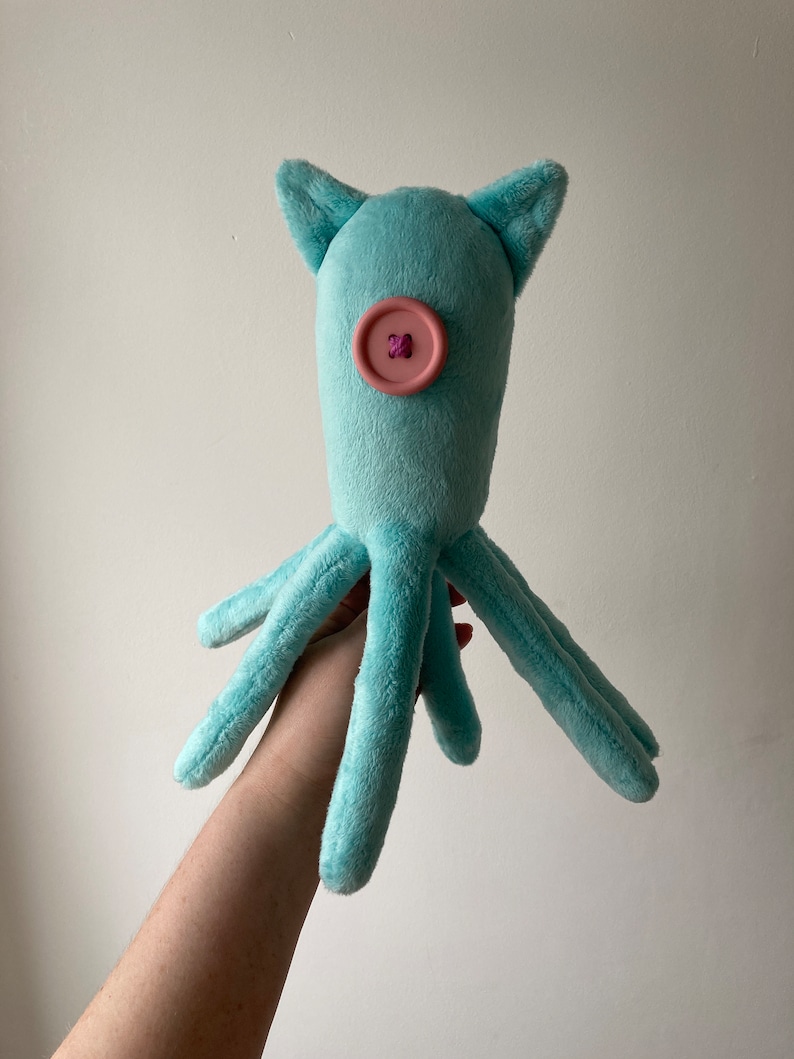 May include: A light blue plush octopus with a pink button for an eye. The octopus has six tentacles and is held in a person's hand.