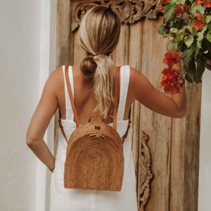 May include: A woman wearing a white dress with a brown woven backpack. The backpack has a rounded shape and a brown leather strap.