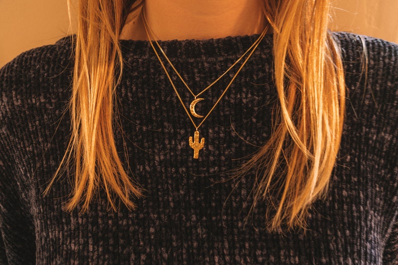 May include: A woman wearing a dark gray sweater with a gold necklace featuring a crescent moon and a cactus pendant.