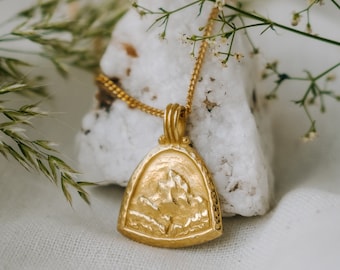 Könnte beinhalten: Eine goldene Halskette mit einem Anhänger in Form einer Bergkette. Der Anhänger ist mit einer strukturierten Oberfläche verziert und hängt vor einem weißen Hintergrund.