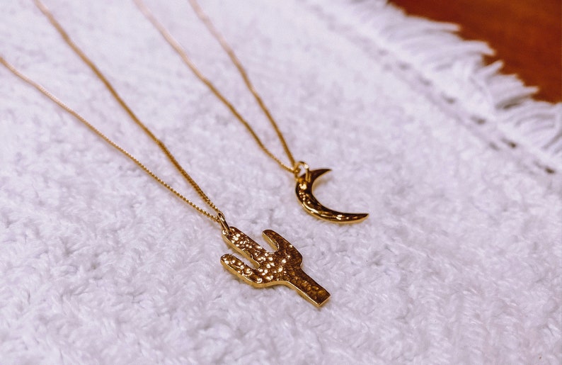 May include: Two gold necklaces with pendants. One pendant is a crescent moon and the other is a cactus. The necklaces are on a white fuzzy surface.