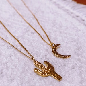 May include: Two gold necklaces with pendants. One pendant is a crescent moon and the other is a cactus. The necklaces are on a white fuzzy surface.
