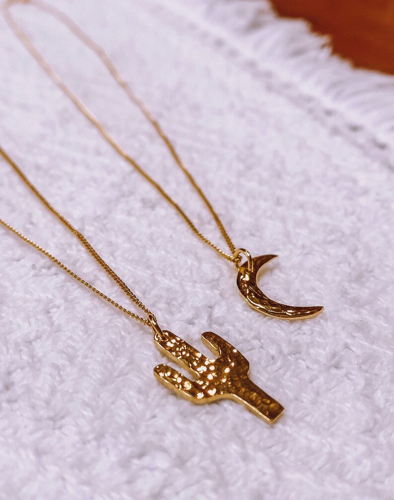 May include: Two gold necklaces with pendants. One pendant is a crescent moon and the other is a cactus. The necklaces are on a white fuzzy surface.