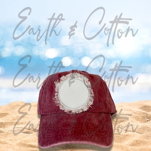 May include: A maroon baseball cap with a distressed, frayed circle design on the front. The cap has a curved brim and a classic six-panel construction. The background features a beach scene with sand and ocean.