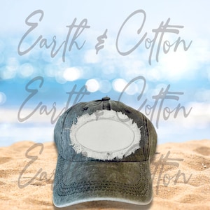 Könnte beinhalten: Eine schwarze, abgenutzte Baseballkappe aus Denim mit einem weißen, ovalen Patch auf der Vorderseite. Die Kappe liegt auf einem Sandstrand.