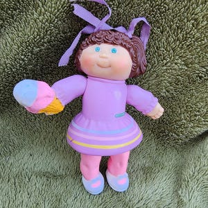 May include: A small Cabbage Patch Kids doll wearing a purple dress with pink and blue stripes, holding a pink, blue, and white ice cream cone. The doll has brown hair, blue eyes, and a pink blush.