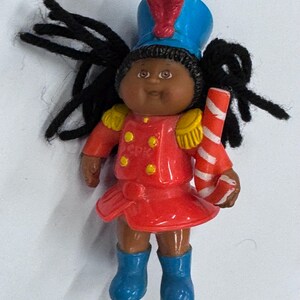 May include: A small doll dressed as a toy soldier. The doll has black yarn hair, brown skin, and is wearing a red jacket, blue boots, and a blue hat with a red plume. It holds a red and white striped candy cane.