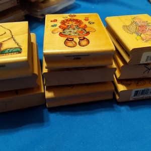 May include: A stack of wooden rubber stamps with various designs, including a girl with flowers, a basket of flowers, and a chick with a sign that says "Treadle or Else!"  The stamps are all in a light wood color and have a white background.