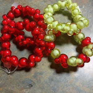 May include: Four beaded napkin rings, two red and two yellow, are arranged on a textured surface. The rings are made of small, round beads that are connected together to form a circle.
