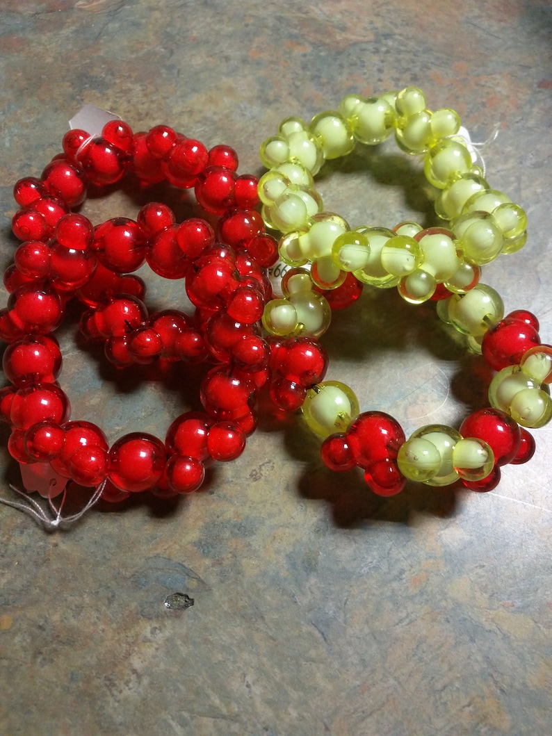 May include: Four beaded napkin rings, two red and two yellow, are arranged on a textured surface. The rings are made of small, round beads that are connected together to form a circle.