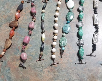 Könnte beinhalten: Sechs Armbänder mit verschiedenen bunten Perlen und Metallakzenten. Die Armbänder haben unterschiedliche Stile und Farben, darunter rosa, grüne, weiße und braune Perlen.