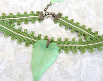 May include: A green choker necklace featuring a woven fabric band with looped edges and a central light green leaf-shaped pendant. The necklace includes a bronze-toned metal clasp and a small matching green leaf charm at the end of the chain.