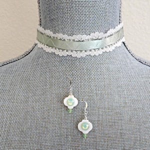 May include: A choker necklace featuring a sage green satin ribbon bordered by white scalloped lace trim, paired with two drop earrings. Each earring consists of a white quatrefoil-shaped charm with a central iridescent bead.