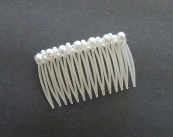 May include: A hair comb featuring a row of white faux pearls of varying sizes attached to the top edge. The comb itself is made of translucent white plastic with fourteen long, tapered teeth designed for securing hair in place.