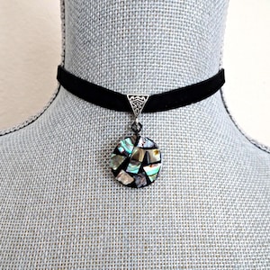May include: A black velvet choker necklace featuring a silver-toned triangular bail and a circular pendant. The pendant is inlaid with iridescent abalone shell fragments in various shapes, creating a mosaic pattern with dark grout lines.