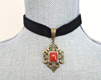 Puede incluir: Un collar tipo gargantilla de terciopelo negro con un colgante de metal en tono latón antiguo. El colgante tiene una piedra roja cuadrada central engastada en un marco de filigrana decorativo con volutas y pequeños detalles de cuentas.