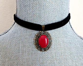Puede incluir: Un collar tipo gargantilla de terciopelo negro con un colgante ovalado central. El colgante tiene una piedra cabujón roja brillante y lisa engastada en un marco de filigrana de metal color bronce envejecido con un borde decorativo festoneado.
