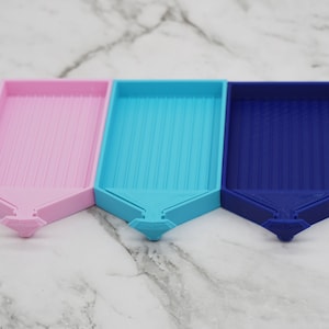 May include: Three diamond painting trays in pink, blue, and dark blue. Each tray has a diamond-shaped base and a raised, ridged surface.