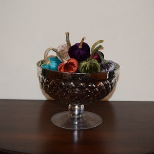 Puede incluir: Un cuenco plateado lleno de calabazas de terciopelo en varios colores, incluyendo turquesa, morado, verde y naranja. El cuenco está sobre una superficie de madera oscura.