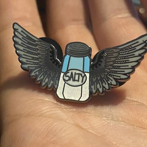 May include: A silver enamel pin with a salt shaker with wings. The salt shaker is blue and white and says "SALTY" on the label.