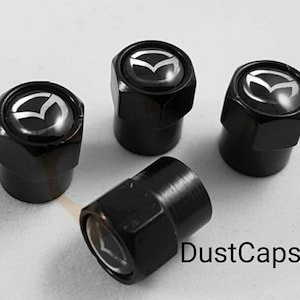 May include: Four black tyre valve stem caps with a silver Mazda logo. The caps are on a white surface.