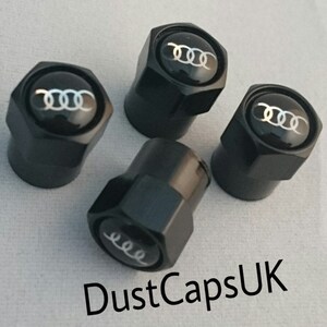 May include: Four black valve stem caps with a silver Audi logo. The caps are hexagonal shaped and have a shiny finish.