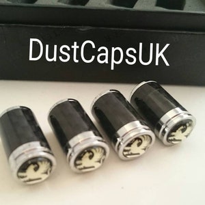 May include: Four black and silver car valve stem caps with a gold Vauxhall logo. The caps are on a white surface.