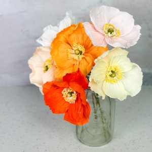 May include: A bouquet of paper flowers in a glass vase. The flowers are in shades of orange, white, and pink. The flowers are arranged in a loose, natural style.