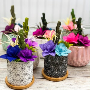 May include: A collection of six small potted plants with paper flowers in various colors, including pink, purple, blue, and green. The pots have a geometric pattern and are arranged on a white wooden surface.