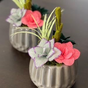 May include: Two gray ceramic pots with a variety of paper flowers. The flowers are pink, white, and green. The pots have a textured surface.