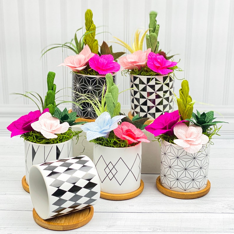 May include: A collection of six black and white geometric patterned planters with colorful paper flowers. The planters are arranged on a white surface.