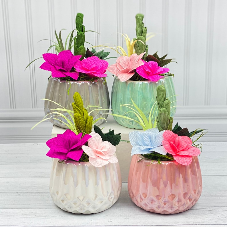 May include: Five ceramic pots with a textured finish, each containing a different arrangement of paper flowers and greenery. The pots are in shades of green and white, and the flowers are in shades of pink, white, and blue.