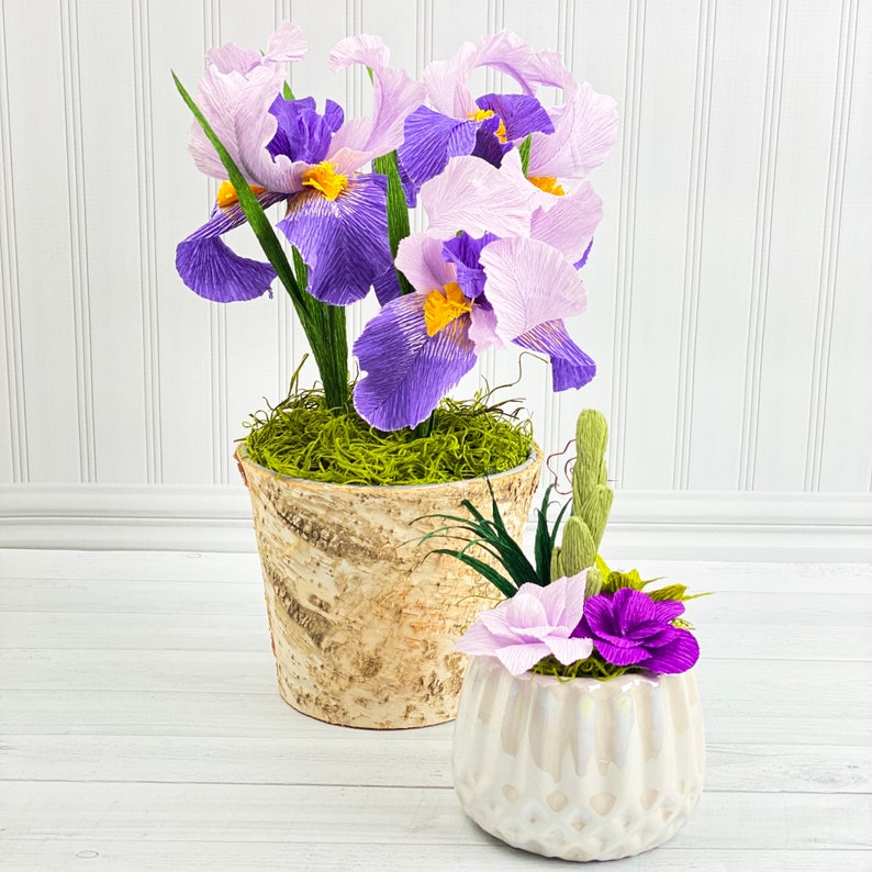May include: Two ceramic pots with paper iris flowers. The larger pot is brown and has a light green moss base. The smaller pot is white with a textured surface. Both pots have purple and light purple iris flowers.