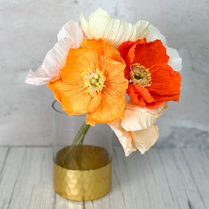 K&ouml;nnte beinhalten: Ein Strau&szlig; aus drei Papierblumen in einer durchsichtigen Glasvase mit goldenem Boden. Die Blumen sind orange, pfirsichfarben und wei&szlig;.
