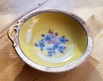 May include: A small, yellow bowl with a gold rim and a floral design. The bowl has two handles and is made of porcelain.