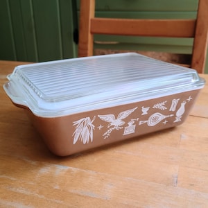 May include: A rectangular copper-colored glass baking dish with a clear, ribbed glass lid. The exterior of the dish features a white printed pattern of an eagle, corn, a coffee grinder, a mortar and pestle, and a lantern along the side.