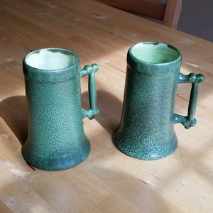 May include: Two green ceramic mugs with handles. The mugs have a speckled glaze and a rounded shape.