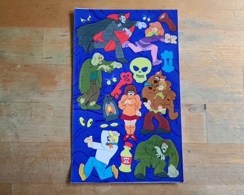 May include: A blue background with a variety of stickers featuring characters from the Scooby-Doo cartoon. The stickers include Scooby-Doo, Shaggy, Daphne, Velma, and a vampire. There are also stickers of a skull, a key, and a lantern.