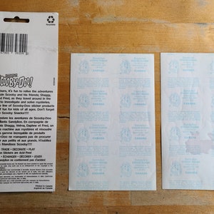 May include: A sheet of Scooby-Doo stickers featuring the Mystery Machine and the Scooby-Doo gang. The stickers are on a white background with blue text. The back of the sheet has instructions for using the stickers in English and French.