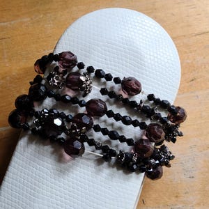 May include: A white sandal with a beaded strap. The strap features multiple strands of black beads, interspersed with dark purple and silver accents. The sandal sits on a wooden surface.