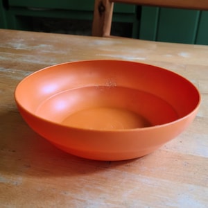 May include: An orange plastic bowl, partially filled with a clear liquid. The bowl is round with a wide rim and sits on a light-colored wooden surface. The bowl's color is a vibrant, solid orange.