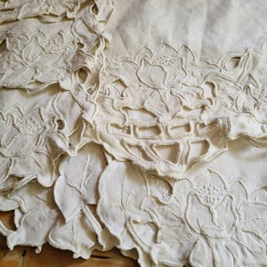 May include: An off-white fabric table runner or placemat featuring intricate cutwork embroidery. The design consists of repeating floral motifs with scalloped edges and openwork details, showcasing detailed stitching on the petals and leaves.
