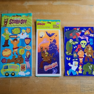 May include: Three packs of Scooby-Doo stickers. The first pack features Scooby and Shaggy with the text "Like,  Whoa!" The second pack features Scooby and Shaggy riding a ghost with the text "Scooby-Doo!" The third pack features Scooby and the gang with the text "Scooby-Doo! Stickers Assortment Collection".