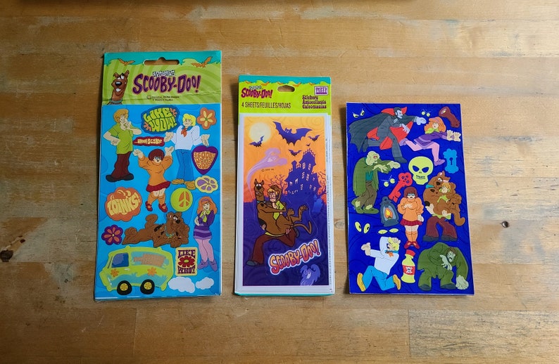 May include: Three packs of Scooby-Doo stickers. The first pack features Scooby and Shaggy with the text "Like,  Whoa!" The second pack features Scooby and Shaggy riding a ghost with the text "Scooby-Doo!" The third pack features Scooby and the gang with the text "Scooby-Doo! Stickers Assortment Collection".