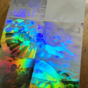 May include: Holographic stickers featuring colorful illustrations of musical instruments, figures, and abstract designs. The stickers have a reflective, iridescent finish, with a rainbow effect. The images are in shades of blue, green, yellow, and purple.
