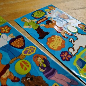 May include: Two sheets of stickers featuring characters from the cartoon Scooby Doo. The stickers are on a blue background and feature various characters, including Scooby Doo, Shaggy, Velma, Daphne, and Fred. The stickers also feature the Mystery Machine van and other iconic elements from the show.