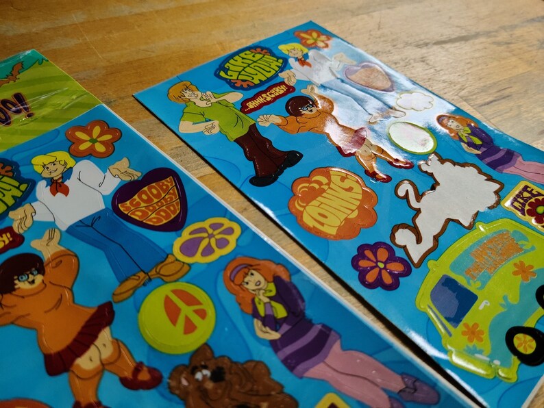 May include: Two sheets of stickers featuring characters from the cartoon Scooby Doo. The stickers are on a blue background and feature various characters, including Scooby Doo, Shaggy, Velma, Daphne, and Fred. The stickers also feature the Mystery Machine van and other iconic elements from the show.