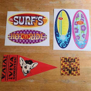 May include: A collection of colorful stickers featuring various designs. Includes oval stickers with "SURF'S UP" and "HOT RIDE" text, surfboard graphics, a red pennant with a dog illustration and text, and a square sticker with "HERE I COME!" text.