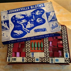 May include: Vintage "Johnnyville Blocks" building set for children aged 4-8. The box features a blue and white illustrated map of a village. The set includes red, pink, and white blocks, along with green tree-shaped pieces.